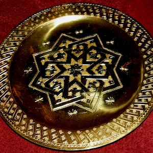May include: A gold-colored metal plate with an intricate, eight-pointed star design in the center. The plate has a decorative border with a repeating pattern.