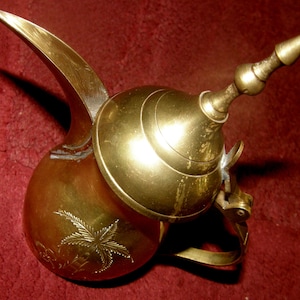May include: A gold brass teapot with a decorative lid and a long spout. The teapot has an engraved design of a palm tree.