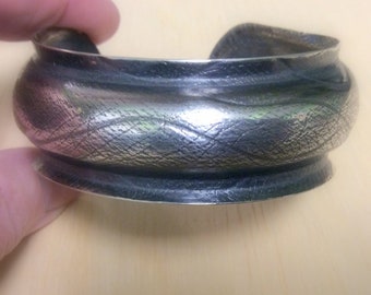 May include: A silver bracelet with a textured surface. The bracelet is curved and has a rounded shape.