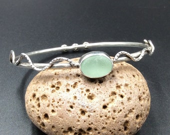 May include: A silver bangle bracelet with a delicate, twisted design and a light green sea glass accent.