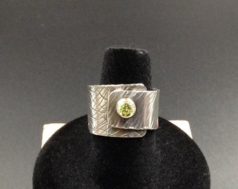 May include: A silver ring with a textured surface and a green gemstone set in the centre. The ring is on a black display stand.