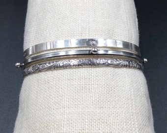 May include: Three silver bracelets stacked on a white fabric background. The top bracelet has a textured surface, the middle bracelet is plain, and the bottom bracelet has a floral design.