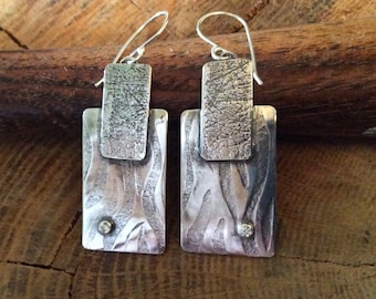 May include: Silver earrings with a textured finish and a small clear stone accent. The earrings are rectangular in shape and have a layered design.