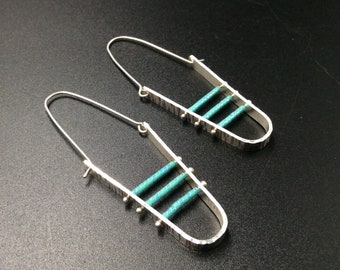 May include: A pair of silver earrings with a geometric design featuring turquoise accents. The earrings are long and dangle, with a textured finish.