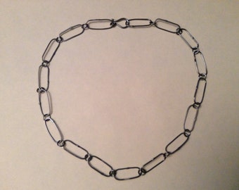 May include: A silver-coloured necklace comprising a series of elongated, oval-shaped links. The links are connected to form a complete circle, with a clasp closure. The necklace is displayed against a neutral background, highlighting its simple, yet elegant design.