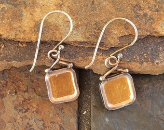May include: A pair of square earrings with gold centres and silver frames. The earrings have a hook design for pierced ears. The background is a textured, brown and grey stone surface.