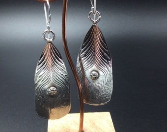 May include: A pair of silver earrings with a textured peacock feather design. The earrings are hanging from a wooden stand.