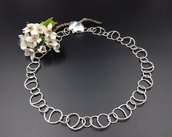 May include: A silver necklace with a chain made of interconnected circles. The necklace is on a black background with a sprig of white flowers.