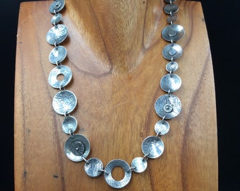 May include: A silver necklace with textured round pendant charms. The necklace is on a wooden mannequin.
