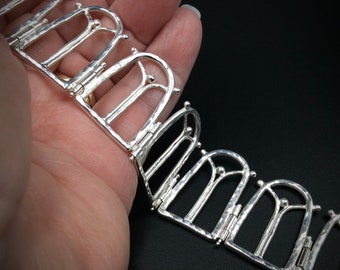 May include: A silver necklace featuring a repeating pattern of arched shapes. The necklace is made up of multiple linked sections, each with a textured surface.