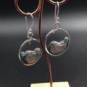 May include: A pair of silver earrings with a textured, circular design and a small turquoise stone. The earrings are hanging from a silver hook.