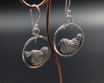 May include: A pair of silver earrings with a textured, circular design and a small turquoise stone. The earrings are hanging from a silver hook.