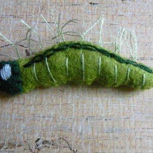 May include: A green felt caterpillar with white stitching and white felt accents. The caterpillar has a segmented body and is curled into a C shape.