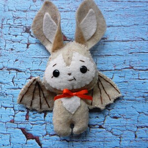 May include: A plush toy bat with brown wings, white fur, and large black eyes. The bat is wearing a red bow and is sitting on a blue wooden surface.