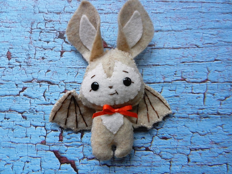 May include: A plush toy bat with brown wings, white fur, and large black eyes. The bat is wearing a red bow and is sitting on a blue wooden surface.