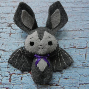 May include: A gray felt bat plush toy with black wings and a purple bow. The bat has black eyes and a stitched smile.