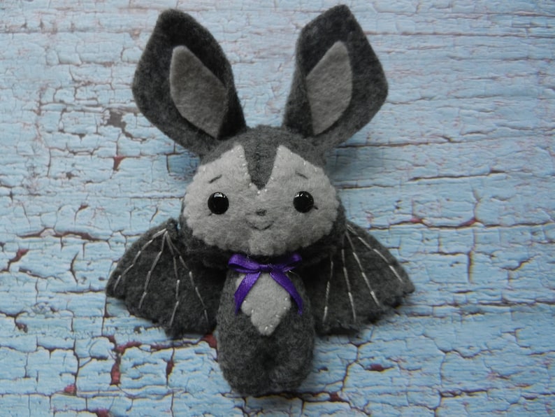 May include: A gray felt bat plush toy with black wings and a purple bow. The bat has black eyes and a stitched smile.