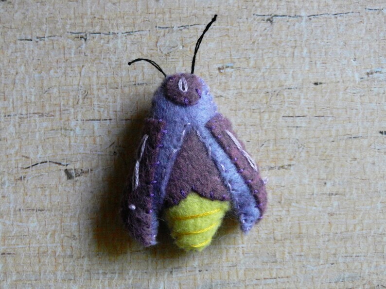 May include: A purple and brown felt moth with yellow wings. The moth has a black thread antenna and is stitched with purple thread.
