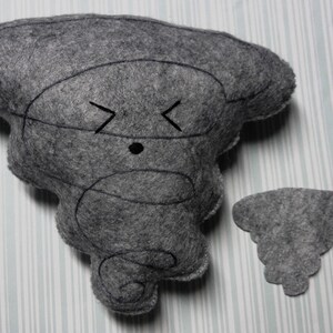 Pu&ograve; includere: Un giocattolo di peluche in feltro grigio a forma di tornado con un viso ricamato nero. Il giocattolo ha una piccola nuvola di feltro grigio attaccata ad esso.
