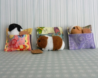 May include: Three plush toys are sleeping in their own colorful fabric beds. The beds are decorated with different patterns and colors. The toys are a black and white penguin, a brown and white guinea pig, and a brown and white dog.