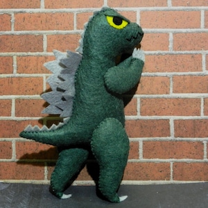 Puede incluir: Juguete monstruo de fieltro verde con una columna vertebral de fieltro gris y un ojo amarillo. El monstruo est&aacute; de pie sobre un fondo de ladrillo.
