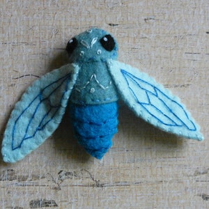 May include: A blue felt insect with embroidered wings and black eyes. The insect has a segmented body and is a handmade craft.