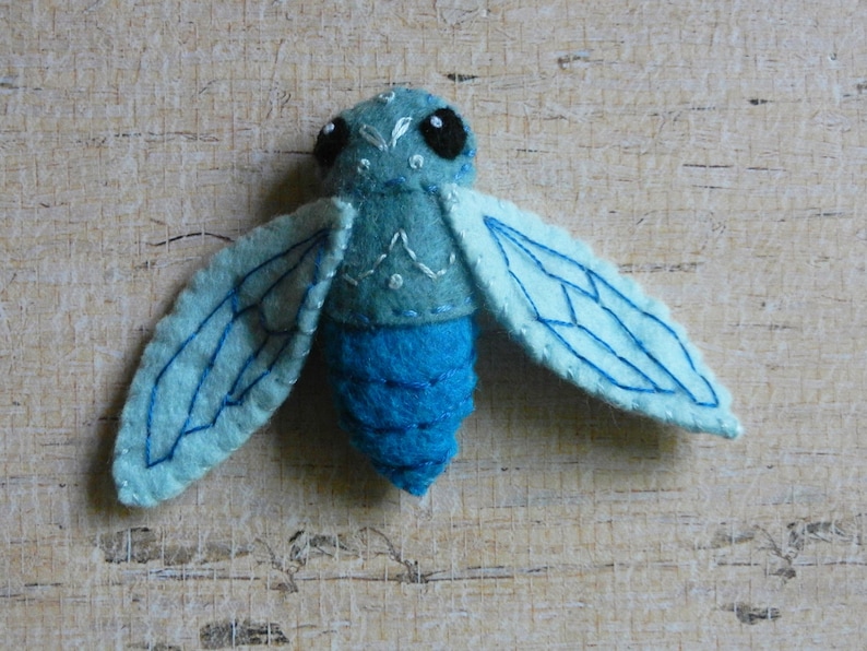 May include: A blue felt insect with embroidered wings and black eyes. The insect has a segmented body and is a handmade craft.