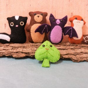 May include: Five felt animal plushies, including a skunk, bear, bat, beaver, and a green leaf, are arranged on a piece of tree bark.
