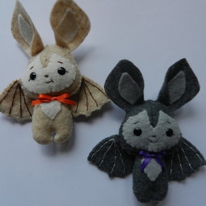 May include: Two felt bat plushies, one brown with orange ribbon and one gray with purple ribbon. Both have embroidered eyes and mouths.