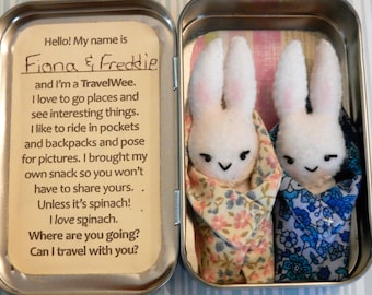 May include: A tin with a handwritten note that reads "Hello! My name is Fiona & Freddie and I'm a TravelWee. I love to go places and see interesting things. I like to ride in pockets and backpacks and pose for pictures. I brought my own snack so you won't have to share yours. Unless it's spinach! I love spinach. Where are you going? Can I travel with you?" Inside the tin are two white felt bunny rabbits with embroidered faces, each wrapped in a patterned fabric. The bunny on the left is wrapped in a floral fabric and the bunny on the right is wrapped in a blue and white floral fabric.