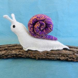 May include: A white felt snail with a purple and gold shell. The snail is sitting on a piece of driftwood.