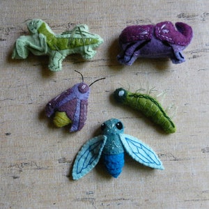 May include: Five felt animal figurines: a green grasshopper, a purple beetle, a green caterpillar, a purple moth, and a blue dragonfly.