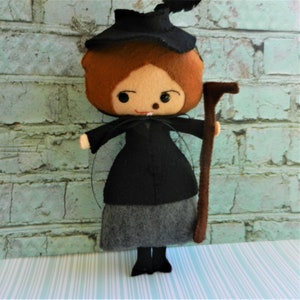 May include: A felt doll dressed as a witch, wearing a black dress, gray skirt, and black hat with a feather. The doll is holding a brown stick.