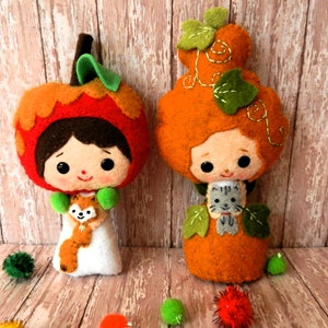 Puede incluir: Dos muñecas de fieltro con forma de calabaza con detalles bordados. Una muñeca sostiene un pequeño zorro de fieltro y la otra sostiene un pequeño gato de fieltro. Las muñecas están de pie sobre una superficie de madera.
