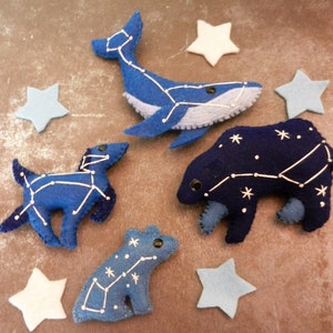 May include: Five felt animal plushies with constellations embroidered on them. The animals are a whale, a dog, a bear, a frog, and a star. The plushies are blue and dark blue with white stitching.
