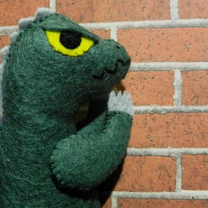 Puede incluir: Juguete monstruo de fieltro verde con un ojo amarillo y p&uacute;as de fieltro gris en la espalda. El monstruo se apoya en una pared de ladrillo.