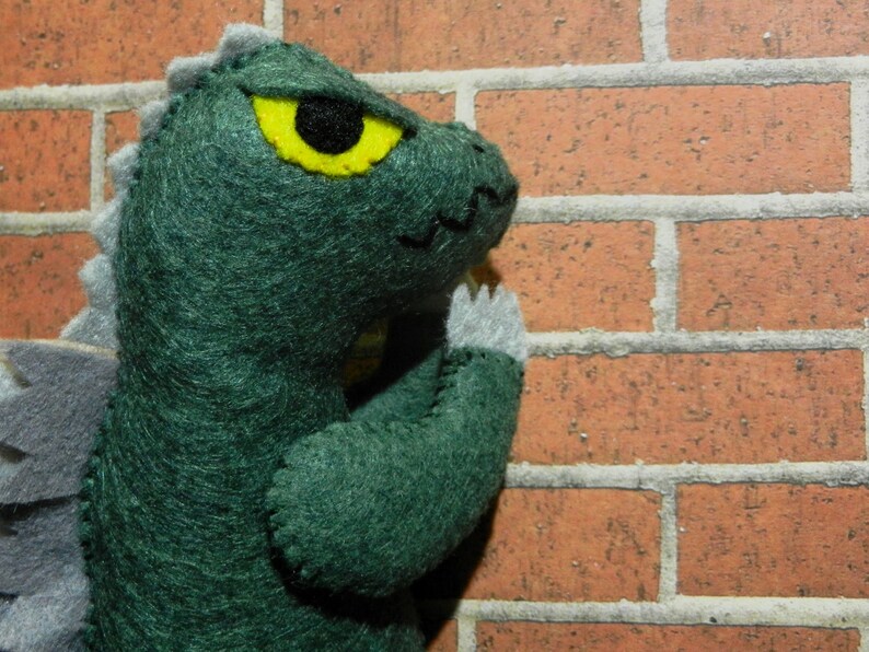 Puede incluir: Juguete monstruo de fieltro verde con un ojo amarillo y p&uacute;as de fieltro gris en la espalda. El monstruo se apoya en una pared de ladrillo.