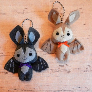 May include: Two felt bat ornaments, one gray with purple ribbon and one brown with orange ribbon. Both bats have black eyes and a smiling mouth.