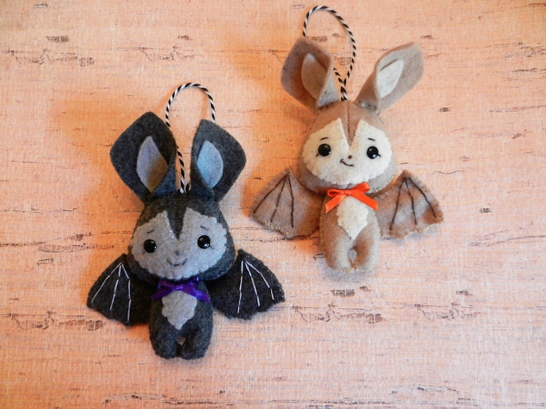 May include: Two felt bat ornaments, one gray with purple ribbon and one brown with orange ribbon. Both bats have black eyes and a smiling mouth.