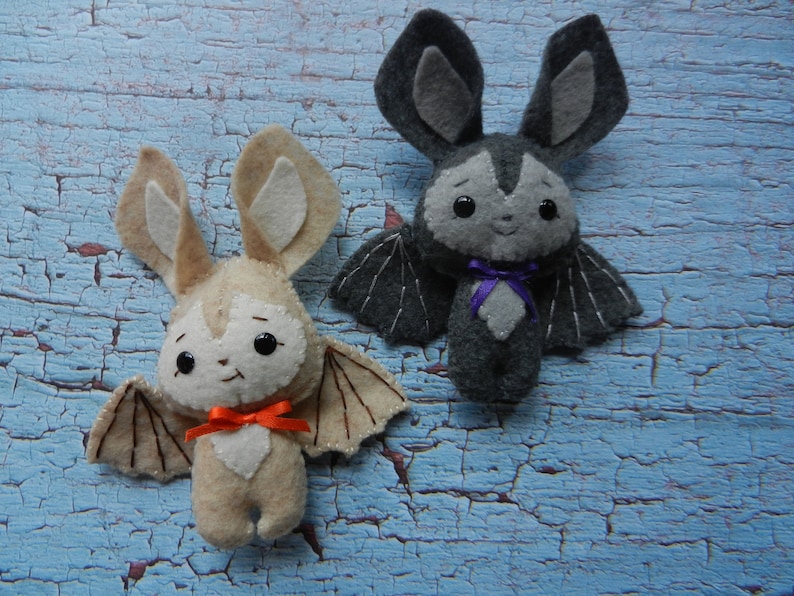 May include: Two felt animal toys, a brown bat with orange ribbon and a gray bat with purple ribbon. Both toys have stitched details and black eyes.