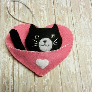 Può includere: Un gatto di feltro nero con tratti del viso bianchi spunta da un cuore di feltro rosa. Il cuore ha un cuore di feltro bianco al centro e un nastro bianco per appenderlo.