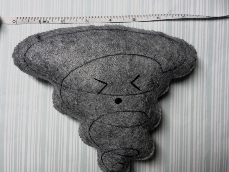 Pu&ograve; includere: Un giocattolo di peluche in feltro grigio a forma di tornado dei cartoni animati con un occhio nero e una smorfia. Il giocattolo &egrave; alto circa 18 cm.