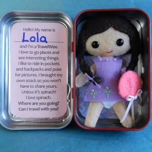 May include: A small, handmade doll named Lola in a purple dress and holding a pink lollipop. The doll is in a tin container with a pink background and text that reads "Hello! My name is Lola and I'm a Travel Wee. I love to go places and see interesting things. I like to ride in pockets and backpacks and pose for pictures. I brought my own snack so you won't have to share yours. Unless it's spinach! I love spinach. Where are you going? Can I travel with you?"