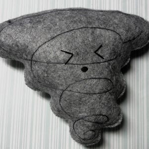Pu&ograve; includere: Un giocattolo di peluche grigio a forma di tornado con un viso da cartone animato. Il giocattolo ha un occhio nero e una bocca semplice con due linee inclinate.