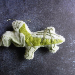May include: A green and white felt praying mantis with embroidered details. The mantis is lying on its side with its legs outstretched.