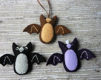 May include: Three felt bat ornaments, one brown, one gray, and one purple. Each bat has a stitched face and wings.