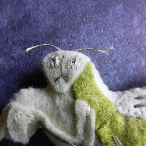 May include: A plush toy in the shape of a praying mantis. The toy is made of white and green felt and has embroidered eyes. The toy is lying on a purple background.