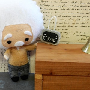 May include: A felt doll of Albert Einstein wearing a brown jacket and gray pants. He is holding a small chalkboard with the equation "E=mc²" written on it. A small gold bell sits on a wooden table behind the doll.