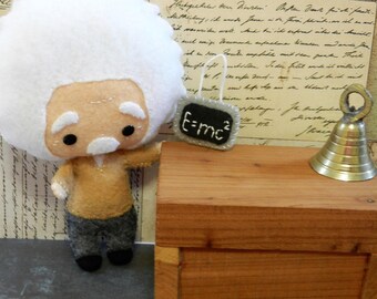 May include: A felt doll of Albert Einstein wearing a brown jacket and gray pants. He is holding a small chalkboard with the equation "E=mc²" written on it. A small gold bell sits on a wooden table behind the doll.