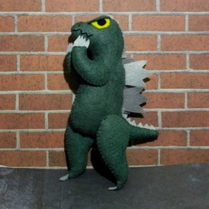 Puede incluir: Juguete monstruo de fieltro verde con un ojo amarillo y una espalda gris puntiaguda. El monstruo est&aacute; de pie sobre una superficie gris frente a una pared de ladrillo.